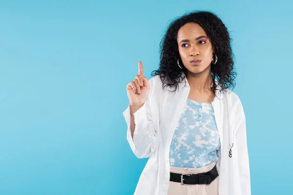 Thoughtful african american woman in white shirt showing idea sign isolated on blue — Stock Photo