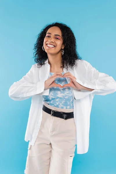 Carefree african american woman in white stylish shirt showing heart sign with hands isolated on blue — Stock Photo