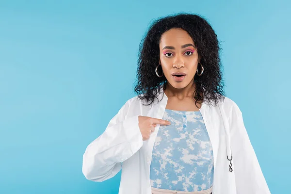 Überraschte Afroamerikanerin im weißen Hemd zeigt mit dem Finger auf sich selbst und blickt vereinzelt in die Kamera auf blauem Grund — Stockfoto