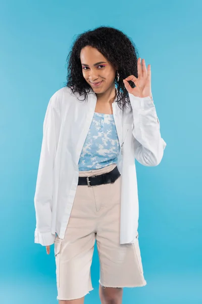 Pleased african american woman in trendy clothes looking at camera and showing okay sign isolated on blue — Stock Photo