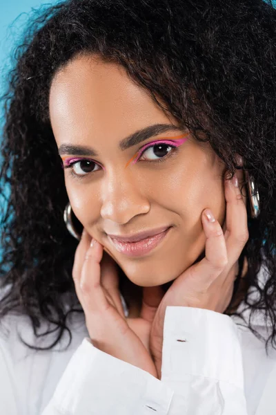 Retrato de encantadora mujer afroamericana con maquillaje y piel perfecta tocando la cara y sonriendo a la cámara aislada en azul - foto de stock