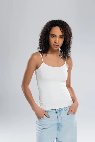 African american woman in tank top posing with hands in pockets of blue jeans isolated on grey — Stock Photo
