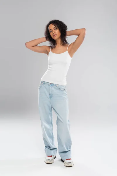 Comprimento total de mulher americana africana elegante em jeans azul posando com as mãos atrás da cabeça no fundo cinza — Fotografia de Stock