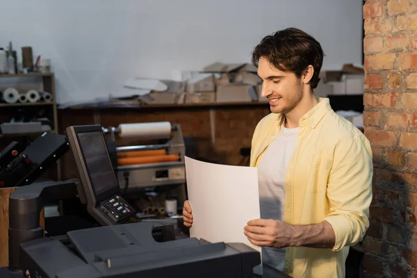 Glücklicher Verleger hält leeres Papier in der Nähe professioneller Ausrüstung im Druckzentrum — Stockfoto