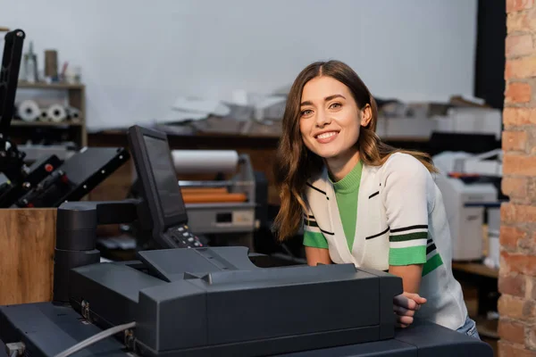 Весела жінка посміхається біля принтера під час роботи в центрі друку — стокове фото
