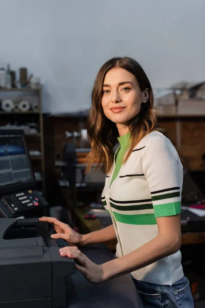 Pretty woman looking at camera while standing near professional equipment in print center — Stock Photo