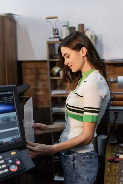Vista lateral da mulher encantadora segurando papéis em branco perto da impressora no centro de cópia — Fotografia de Stock