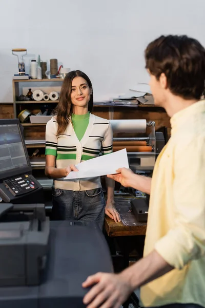 Happy publisher giving blank paper to colleague on blurred foreground in print center — Stock Photo