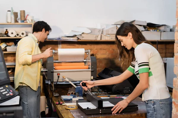 Tipógrafo alegre usando aparador de papel perto colega ao lado de plotter impressão profissional — Fotografia de Stock