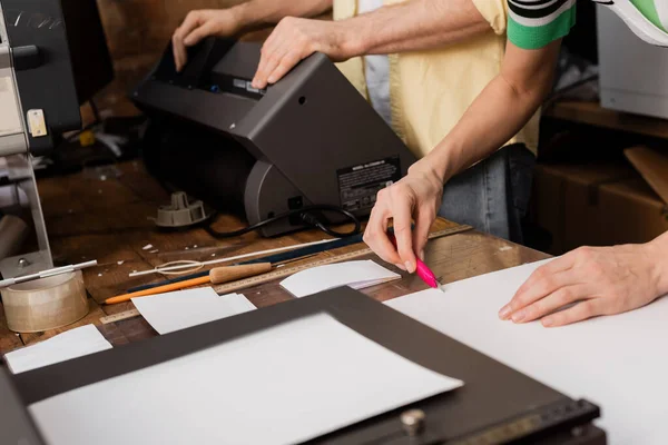 Vista recortada del tipógrafo cortando papel con cuchillo cerca de colega en centro de impresión - foto de stock