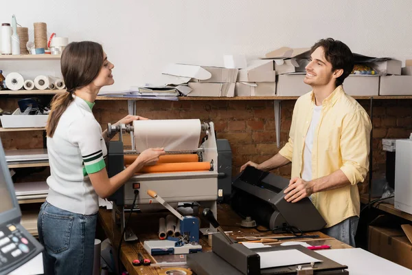 Fröhliche Frau beim Papierziehen mit Druckplotter neben glücklichem Kollegen — Stockfoto