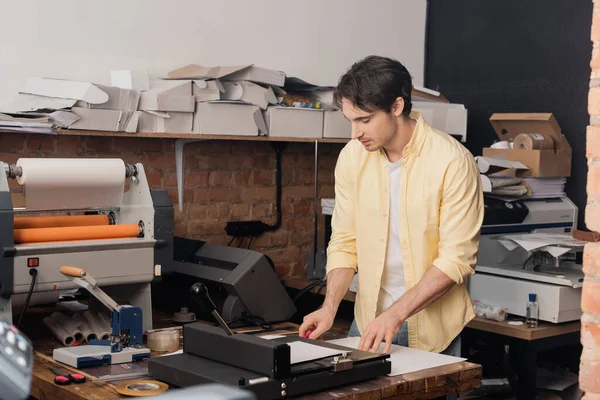 Gut aussehender Typograf schneidet Papier mit Messer in der Nähe von Papierschneider im Druckzentrum — Stockfoto