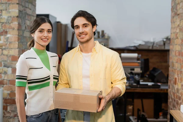 Hombre feliz sosteniendo caja de cartón cerca de mujer alegre en el centro de impresión - foto de stock