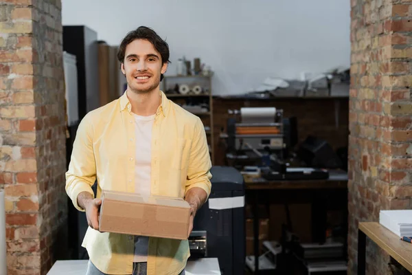 Homem feliz segurando caixa de papelão e olhando para a câmera no centro de impressão — Fotografia de Stock