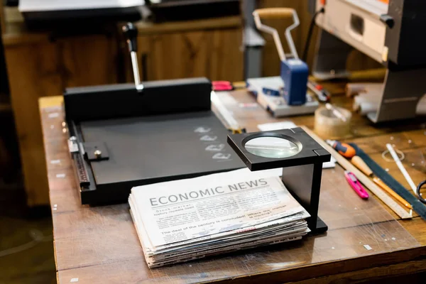 Magnifying glass over newspapers with economic news and professional paper trimmer machine — Stock Photo