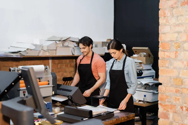 Cheerful man in apron working with printer next to pretty colleague in print center — Stock Photo