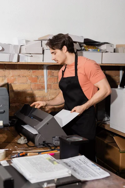 Hombre guapo en delantal sosteniendo papel cerca de la impresora y periódicos en el centro de impresión - foto de stock