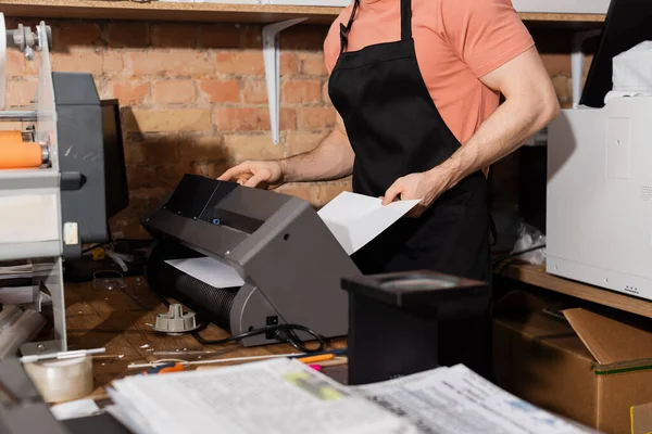 Vista cortada do trabalhador em avental segurando papel perto de impressora e jornais frescos no centro de impressão — Fotografia de Stock