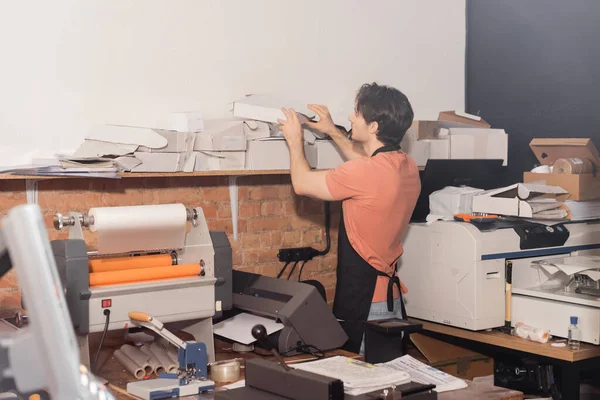 Jovem tipógrafo em avental atingindo caixas de papelão dobradas ao lado de equipamentos no centro de impressão — Fotografia de Stock
