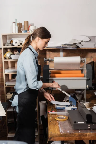 Татуированная женщина в фартуке, работающая с штамповочной машиной для бумаги — стоковое фото