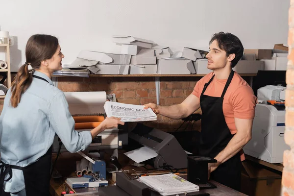 Good looking man giving newspaper with economic news lettering to pretty colleague in print center — Stock Photo