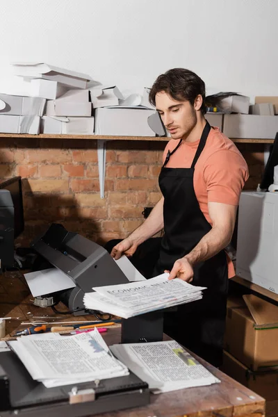 Tipógrafo guapo en delantal con periódicos impresos en la sala de fotocopias - foto de stock
