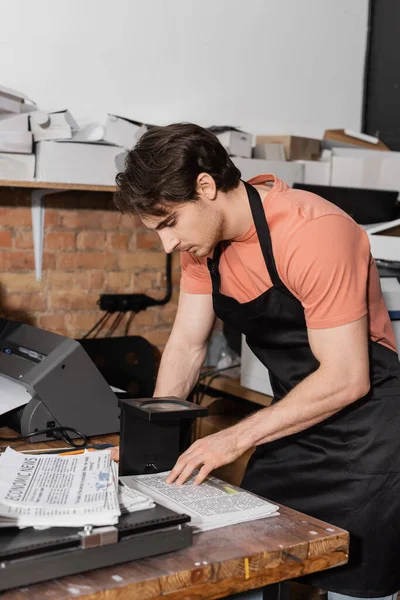 Typographer in apron checking quality of newspaper through magnifying glass in print center — Stock Photo