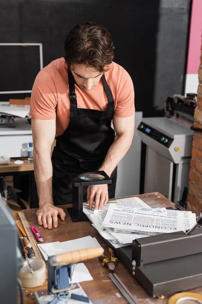 Worker in apron checking quality of newspaper through magnifying glass in print center — Stock Photo