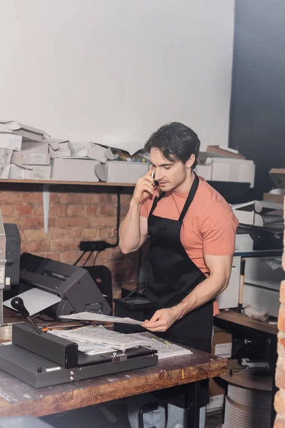 Junger Typograph in Schürze hält Zeitung in der Hand und spricht im Druckzentrum auf dem Smartphone — Stockfoto