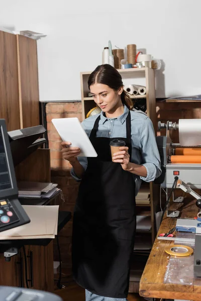 Tipógrafo bonito em avental segurando copo de papel e usando tablet digital no centro de impressão — Fotografia de Stock