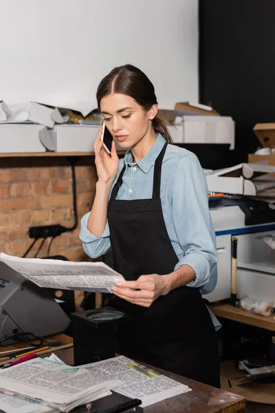 Typographer in apron looking at newspaper while having phone call in print center — Stock Photo
