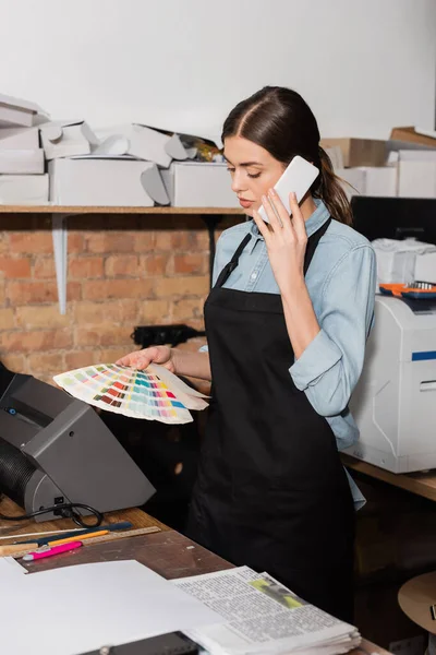 Typographer in apron looking at color samples while having phone call in print center — Stock Photo
