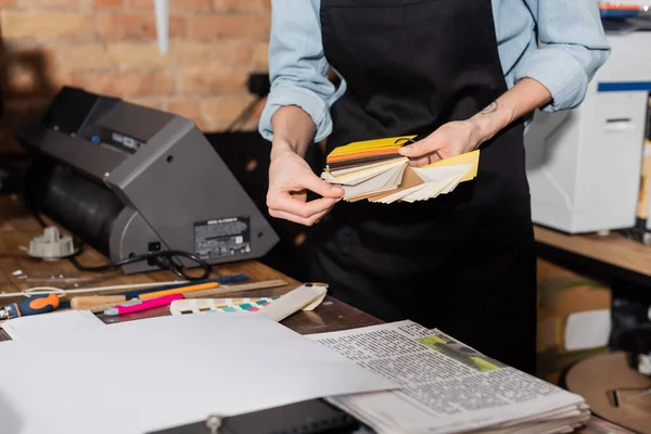 Cropped view of tattooed typographer in apron choosing color samples near newspapers in print center — Stock Photo