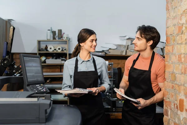 Happy typographers in aprons looking at each other while holding newspapers in print center — Stock Photo