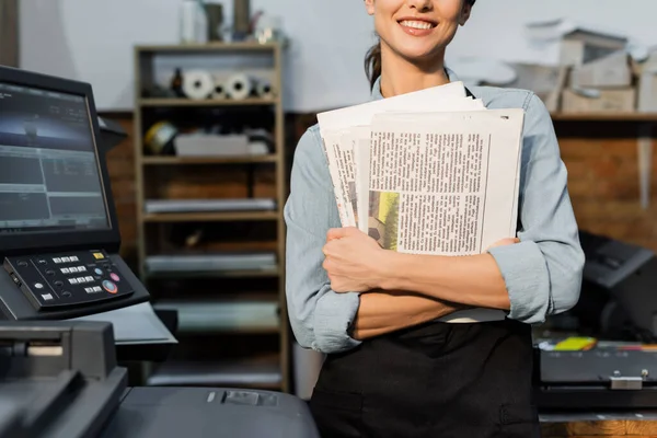 Partial view of positive typographer in apron holding printed newspapers — Stock Photo