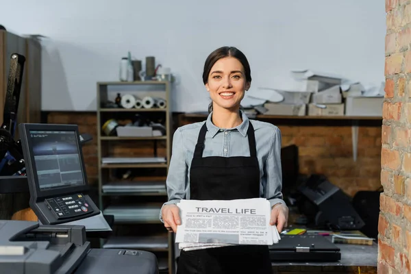 Pretty and positive typographer in apron holding freshly printed travel life newspapers — Stock Photo