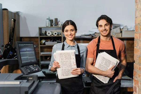 Positive Typographen in Schürzen lächeln, während sie gedruckte Zeitungen in der Hand halten — Stockfoto