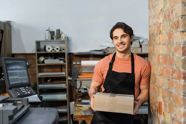 Homem alegre no avental segurando caixa de papelão e sorrindo no centro de impressão — Fotografia de Stock