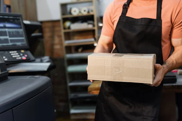 Обрізаний вид працівника в фартусі, що тримає картонну коробку в центрі друку — стокове фото