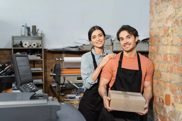 Cheerful typographer leaning on happy colleague with carton box — Stock Photo