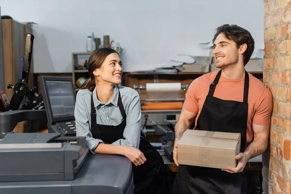 Happy woman in apron leaning on printer and looking at colleague with carton box in print center — Stock Photo