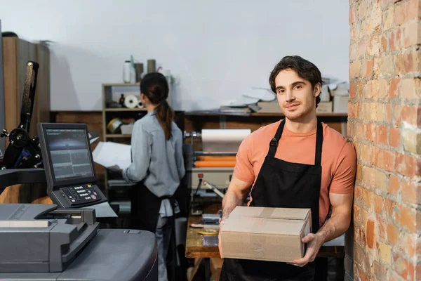 Trabalhador alegre no avental segurando caixa de papelão perto colega feminino no centro de impressão — Fotografia de Stock