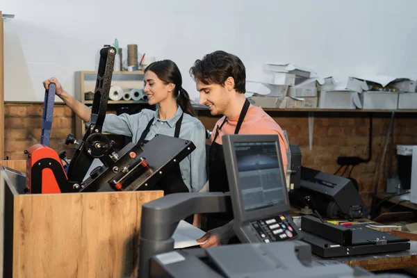 Веселый мужчина и женщина в фартуках с помощью профессионального плоттера печати во время совместной работы в типографии — стоковое фото