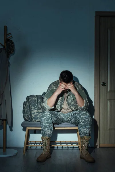 Soldado en uniforme que sufre de trastorno de estrés postraumático mientras está sentado en el pasillo en casa por la noche - foto de stock