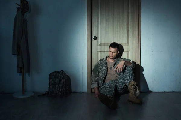 Veterano militar frustrado en uniforme sentado en el piso cerca de la puerta en el pasillo por la noche - foto de stock