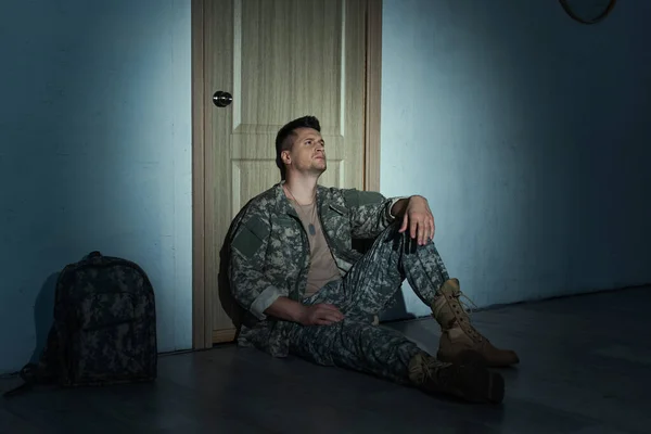 Depressed soldier sitting on floor near backpack and door in hallway at night — Stock Photo