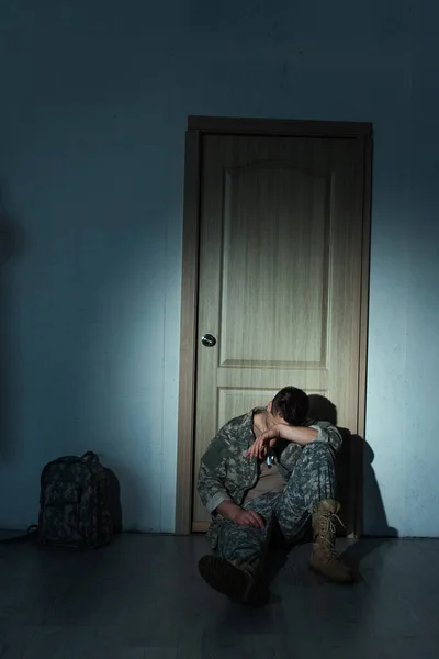 Soldado con trastorno mental sentado cerca de la mochila y la puerta en casa por la noche - foto de stock