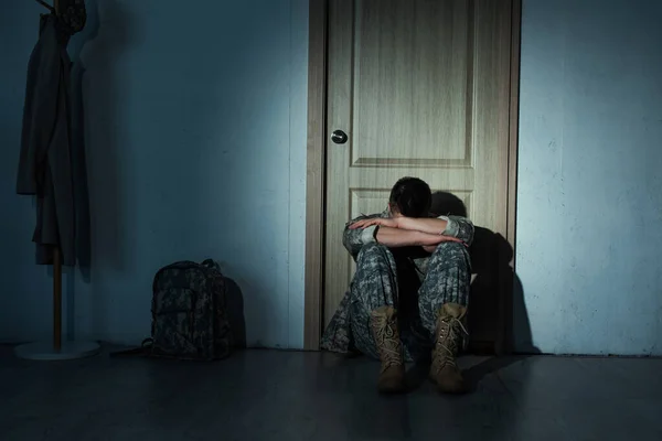 Одинокий солдат в форме сидит ночью возле рюкзака и двери дома — стоковое фото