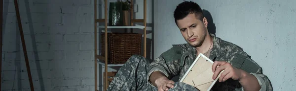 Soldier in uniform taking photo frame from backpack while suffering from emotional distress at home at night, banner — Stock Photo