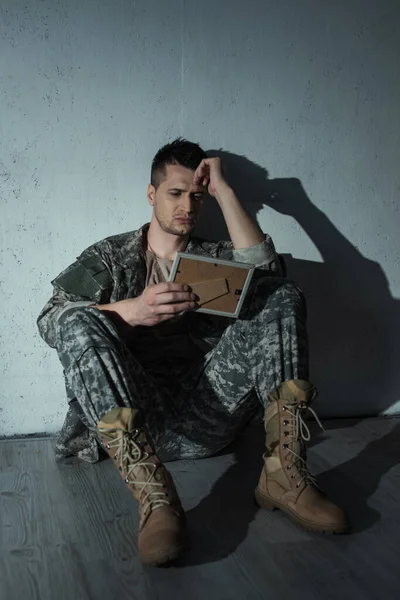 Розчарований військовий ветеран дивиться на фоторамку, сидячи на підлозі вдома вночі — стокове фото